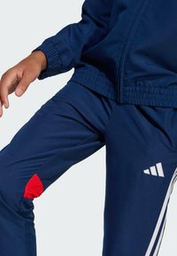 Blå atletiska träningsoverallsbyxor med vita ränder och Adidas-logotyp, med en röd detalj vid knäområdet, tillverkade av ett slätt och flexibelt material.