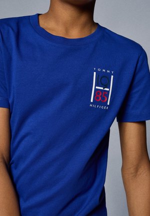 Person trägt ein blaues T-Shirt mit dem Aufdruck "Tommy Hilfiger 1985" in Weiß, Dunkelblau und Rot auf der oberen linken Brustseite.