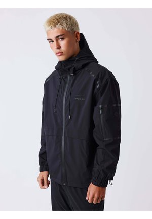 Jeune homme aux cheveux blonds courts portant une veste noire à capuche avec fermeture éclair et poignets élastiques, debout devant un fond clair uni.
