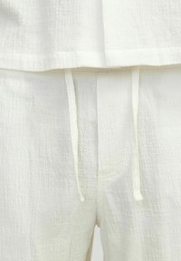 Ensemble de vêtements d'intérieur blancs en tissu texturé et froissé. Le haut a un décolleté ouvert, tandis que le pantalon présente des détails de taille à cordon.