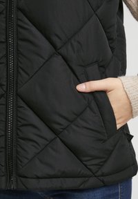 Czarna pikowana kamizelka z zamkiem błyskawicznym i boczną kieszenią. Charakteryzuje się teksturowaną powierzchnią i prostym dołem, zestawiona z dzianinowym swetrem pod spodem.