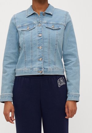 Personne portant une veste en jean bleu clair avec des boutons argentés et un pantalon bleu marine avec le logo blanc GAP sur la partie supérieure de la cuisse gauche.