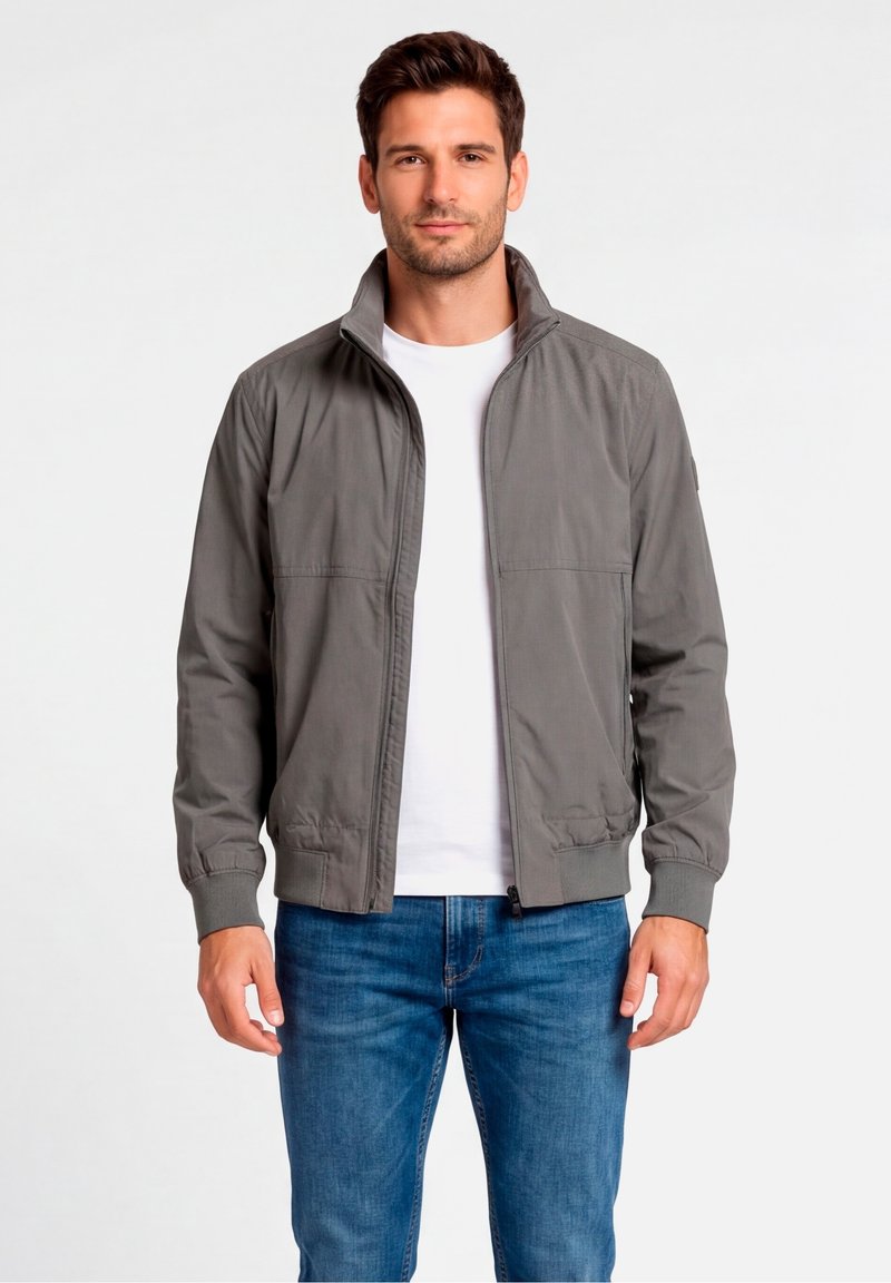 Veste bomber grise avec col montant, fermeture éclair et poignets élastiques, confectionnée dans un matériau léger ; portée par-dessus un t-shirt blanc.