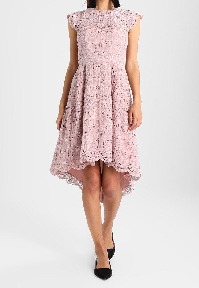 Vaaleanpunainen pitsi mekko, jossa on lyhyet hihattomat hihat, monimutkaiset kuviot, tiukka yläosa ja aallotettu helma, yhdistettynä mustiin korkokenkiin.