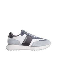 Zapatilla gris y blanca que presenta materiales de ante y cuero, con una suela texturizada, logo en el lateral y un diseño de perfil bajo.