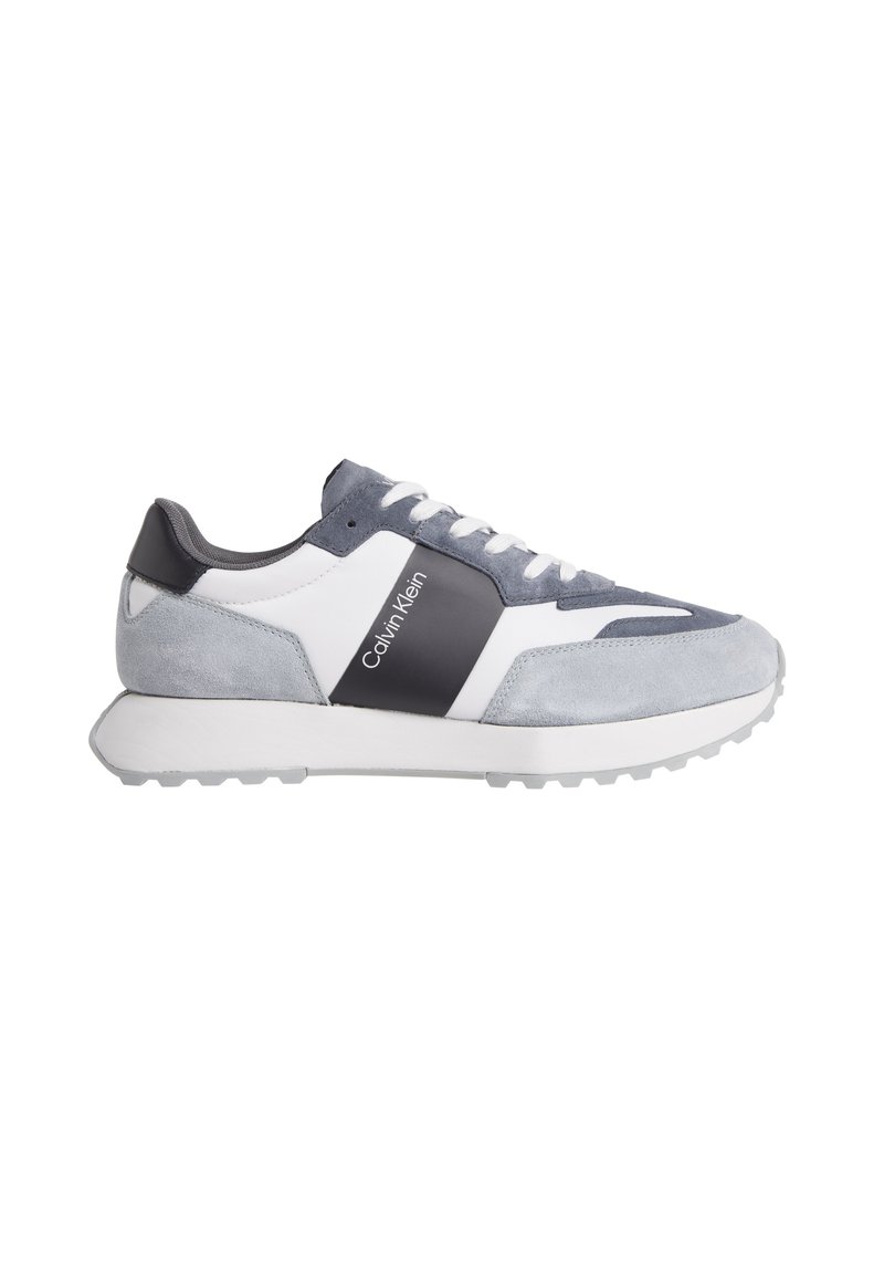 Zapatilla gris y blanca que presenta materiales de ante y cuero, con una suela texturizada, logo en el lateral y un diseño de perfil bajo.