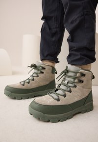 Grüne und beige High-Top-Stiefel aus Wildleder und Gummi. Mit strukturierter Oberseite, kontrastierenden Schnürsenkeln und robustem Profil.