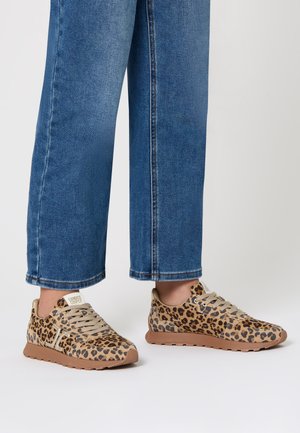 Piedi con jeans blu e sneakers a stampa leopardo con suole beige e lacci color beige su uno sfondo bianco.