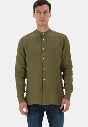 Uomo che indossa una camicia verde oliva con collo alla coreana e jeans blu scuro, in piedi davanti a uno sfondo semplice.