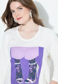 Valkoinen t-paita, jossa on graafinen kuva laventelinvärisistä koroista ja kuvioiduista sukista, joissa on kukkakuviot. Kangas näyttää pehmeältä ja kevyeltä.