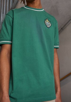 Grünes Baumwoll-T-Shirt mit kurzen Ärmeln, weißem Saum und einem bestickten Emblem auf der Brust, mit glatter Textur und klassischem Schnitt.