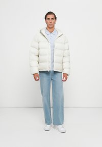 Homme se tenant contre un fond blanc, portant une doudoune blanche, un polo bleu clair, un jean ample et des baskets blanches.