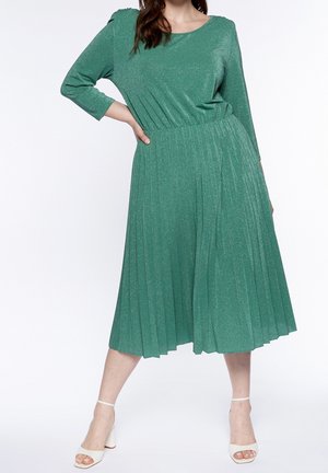 Femme portant une robe midi plissée vert scintillant à manches trois-quarts et des talons blancs à bride à la cheville, debout avec une main sur la hanche.