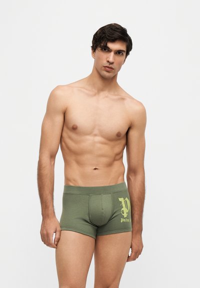 Homme torse nu avec cheveux courts et foncés portant un boxer vert avec un logo jaune, debout devant un fond clair uni.