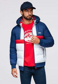 Padded jacket w górnej części w kolorze granatowym i dolnej białej, z czerwoną poziomą paskiem. Zawiera kaptur i zamek błyskawiczny z przodu.