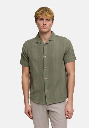Homme blond portant une chemise olive à manches courtes boutonnée et un pantalon beige clair, debout devant un fond uni.
