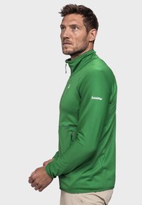 Grüne Fleecejacke mit Reißverschluss und hohem Kragen, mit strukturiertem Muster, einer Seitentasche und einem Logo am Ärmel.