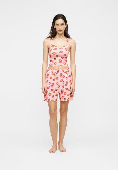 Femme debout pieds nus, portant un haut sans manches rose et un short avec un motif de lèvres rouges sur un fond blanc uni.
