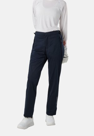 Kvinde iført hvid langærmet top, marineblå bukser med lynlås, hvide sneakers og en hvid handske, står med hænderne på ryggen.