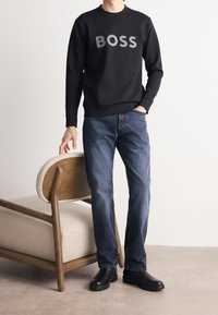 Sudadera negra con la palabra "BOSS" en letras grises en negrita, combinada con unos vaqueros azules y botas de tobillo negras, de pie al lado de una silla de madera.