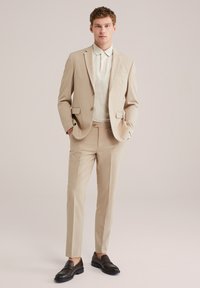 Hombre vestido con un traje beige, camiseta tipo polo color crema y mocasines marrón oscuro, de pie con las manos en los bolsillos frente a un fondo liso.