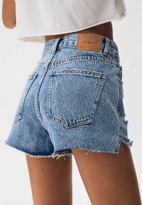 Primer plano de una persona que lleva pantalones cortos de mezclilla azul claro con los bordes desgastados y una etiqueta marrón de Pull&Bear en la cintura.