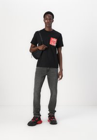 Svart t-skjorte med rødt blomsterlomme-design, grå jeans og svarte sneakers med røde accenter. Modellen holder en liten svart veske.