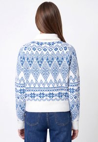 Balta megzta palaidinė su mėlynais geometriniais ir snaigių raštais, su briaunotu apykakliu ir rankogaliais, trumpesnio ilgio. Dėvima su mėlynomis džinsinėmis kelnėmis.