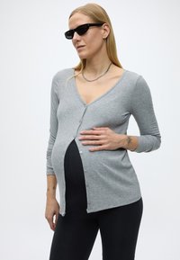 Femeie însărcinată purtând ochelari de soare negri, cardigan gri cu nasturi parțial deschis peste colanți negri, cu mâna sprijinită pe burtă, pe un fundal simplu.