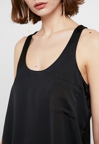 Canotta nera in satin con scollatura a barca e una piccola tasca applicata sul lato sinistro. La texture appare liscia e morbida.