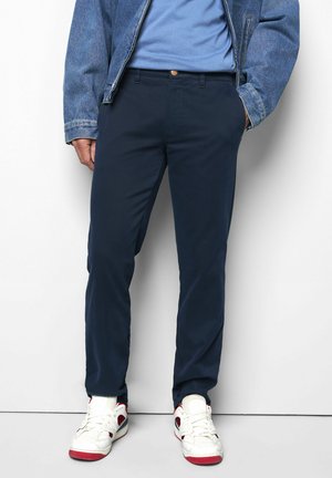Man wearing navy pants, white sneakers with red accents, blue t-shirt, and denim jacket with hand in pocket against a white wall.