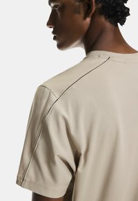 Bežové tričko s krátkymi rukávmi, s hladkou textúrou a čiernymi švami pozdĺž ramenných švov.