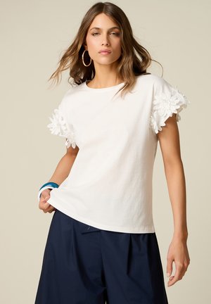 Donna che indossa una camicia bianca a maniche corte con dettagli in pizzo floreale sulle maniche, pantaloni larghi blu navy, orecchini a cerchio dorati e braccialetto blu.
