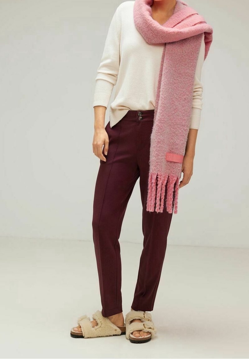 Personne portant un pull crème, un pantalon bordeaux, des sandales beiges duveteuses et une longue écharpe tricotée rose drapée sur une épaule.