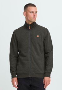 Dark gray zip-up sweatshirt with a high collar, textured fabric, and a small logo patch on the chest. Features zippered closure and side pockets.