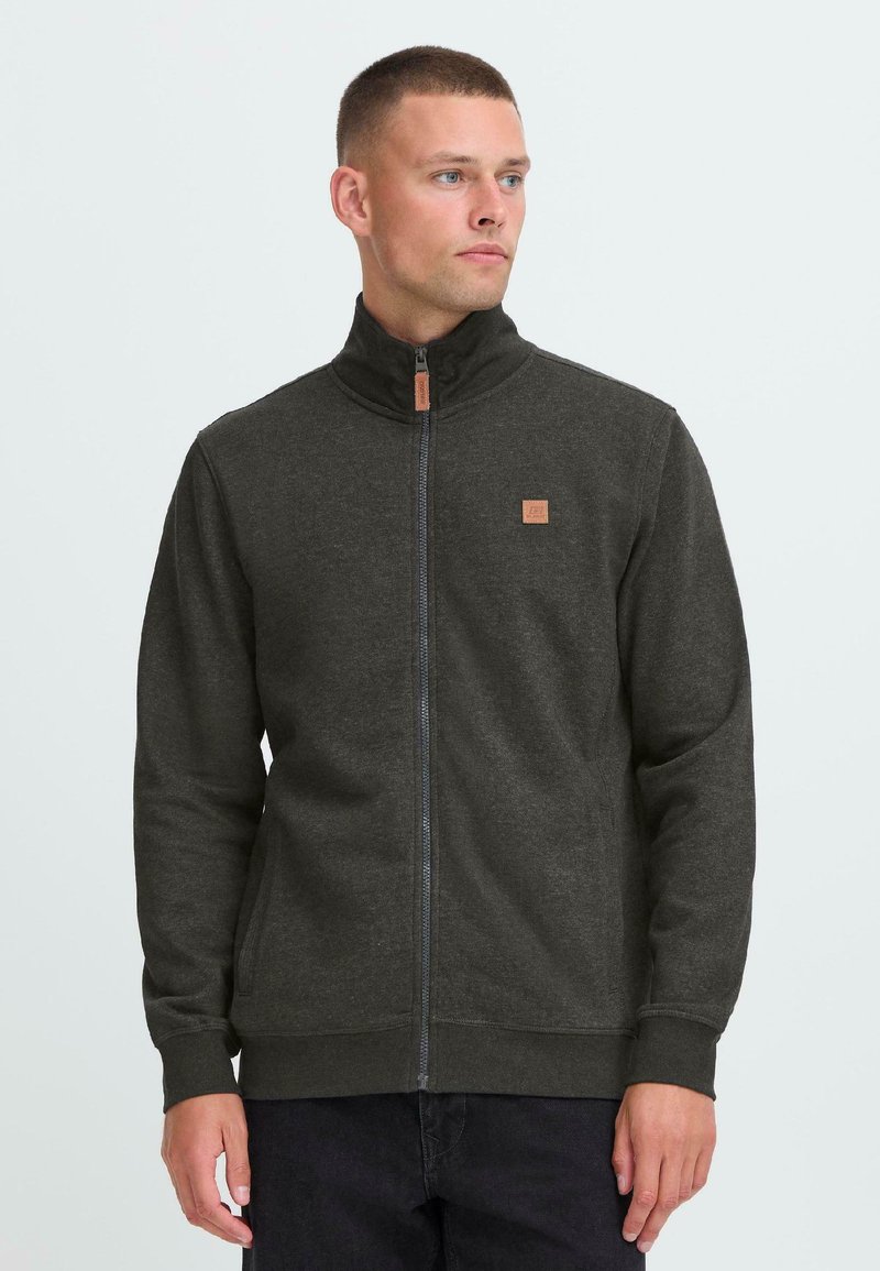 Dark gray zip-up sweatshirt with a high collar, textured fabric, and a small logo patch on the chest. Features zippered closure and side pockets.