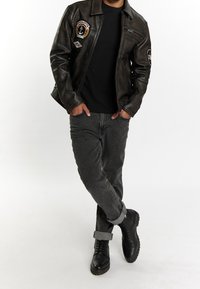 Ruskea nahkainen moottoripyörätakki, jossa on merkkejä, musta t-paita, harmaat farkut ja mustat saappaat käännetyillä resoreilla. Rentoa, kerrospukeutumiseen sopivaa asua.