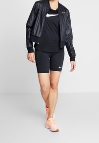 Personne portant une veste de sport noire Nike, un short noir Nike, un t-shirt noir Nike et des chaussures de course couleur pêche, se tenant sur une surface grise uniforme.