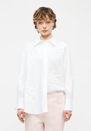 Nuori henkilö, jolla on lyhyet hiukset, valkoinen napillinen paita, jossa on kukkaistikkaus, ja vaaleanpunaiset housut yksiväristä taustaa vasten.