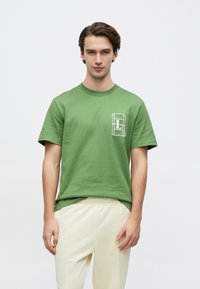 Πράσινο βαμβακερό μπλουζάκι με στρογγυλή λαιμόκοψη, κοντά μανίκια και λευκό γραφικό λογότυπο στο αριστερό στήθος. Μινιμαλιστικός σχεδιασμός.