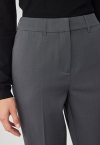 Pantaloni gri, croiți pe măsură, cu o textură netedă, față plată, băiere pentru curea și buzunare laterale. Materialul pare a fi ușor și rezistent la cute.