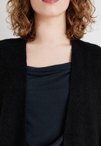 Cardigan negru tricotat cu un model texturat, purtat peste un top drapat de culoare albastru marin. Țesătura pare moale și are o croială lejeră.