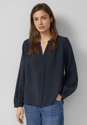 Femme aux longs cheveux bruns portant un chemisier ample bleu marine foncé avec un col en V et un jean bleu, debout devant un fond gris clair uni.