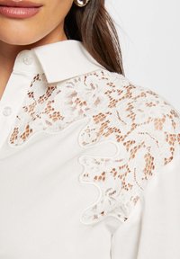 Camicia bianca con colletto classico, caratterizzata da dettagli in pizzo intricati sulle spalle e sulle maniche, che aggiungono texture e interesse visivo.
