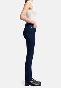 Jeans oscuros de corte slim con una textura suave, que cuentan con una cintura alta, cinco bolsillos y acentos en dorado. Se llevan con mocasines negros.