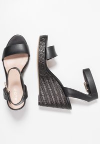 Černé klínové sandály s tkaným texturovaným černým platformou, koženými popruhy a přezkou z růžového zlata na pásku kolem kotníku.