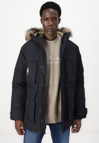 Parka nera con cappuccio foderato in pelliccia sintetica, dotata di numerose tasche frontali, chiusura a zip e una morbida fodera interna sopra una felpa beige.