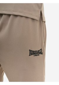 Beežid soonikkoes kangast püksid, millel on musta värvi tikitud "Lonsdale London" logo ülemisel reiel, kombineeritud sobiva ülaäärisega.