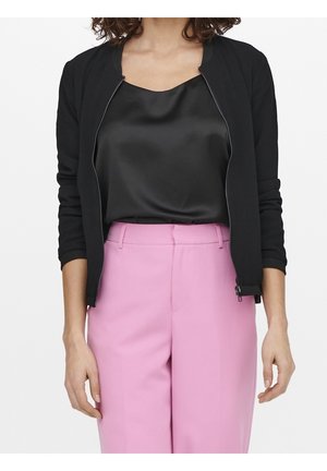 Frau trägt ein schwarzes Satin-Camisole, eine schwarze Reißverschlussjacke mit gerafften Ärmeln und hoch taillierte rosa maßgeschneiderte Hose, steht mit entspannten Armen.
