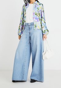Blusa floreale a maniche lunghe, tessuto azzurro chiaro; abbinata a jeans a gamba larga in un lavaggio chiaro; tiene in mano una piccola borsa bianca; scarpe nere con pois bianchi.